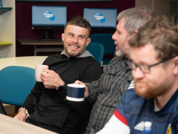 A group of men gathered round a table in the library, drinking hot drinks and smiling.