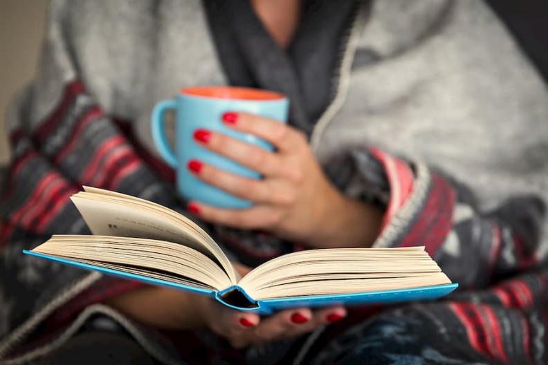 Close-up of a person reading an open book, holding a mug in the other hand.