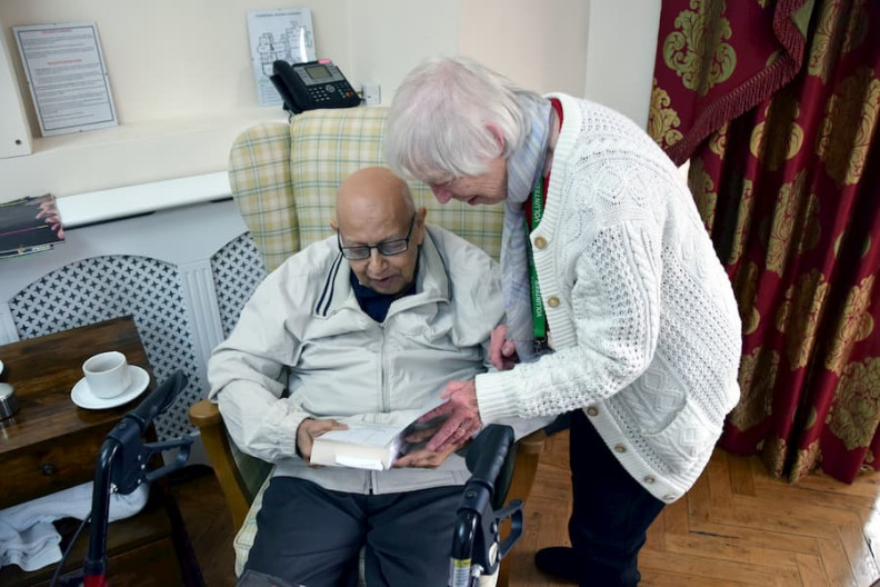 An elderly man sits in a chair holding a book next to a volunteer