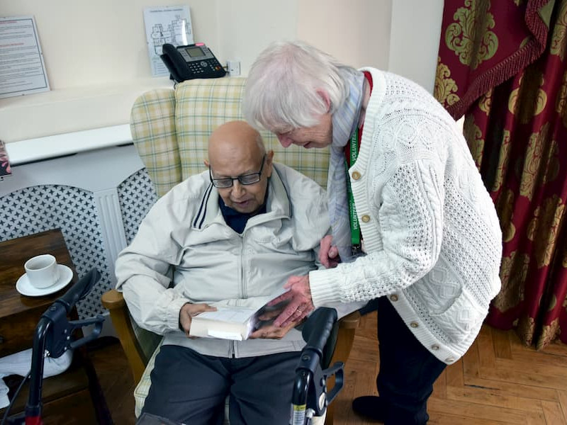 An elderly man sits in a chair holding a book next to a volunteer