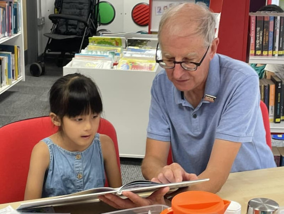 Martyn, a Suffolk Libraries volunteer, sits and reads with a young girl at Saxmundham Library.
