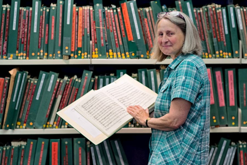A woman holding a binder of sheet music standing in front of shelves full of more binders.
